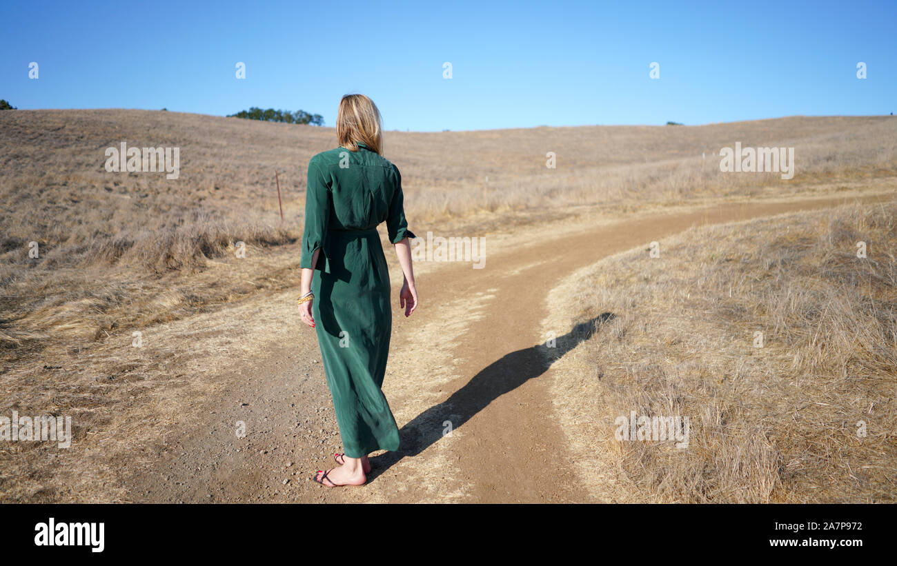 young blonde woman in long green dress standing facing away from camera casting a long shadow upon dirt path in a dry ground, country setting Stock Photo