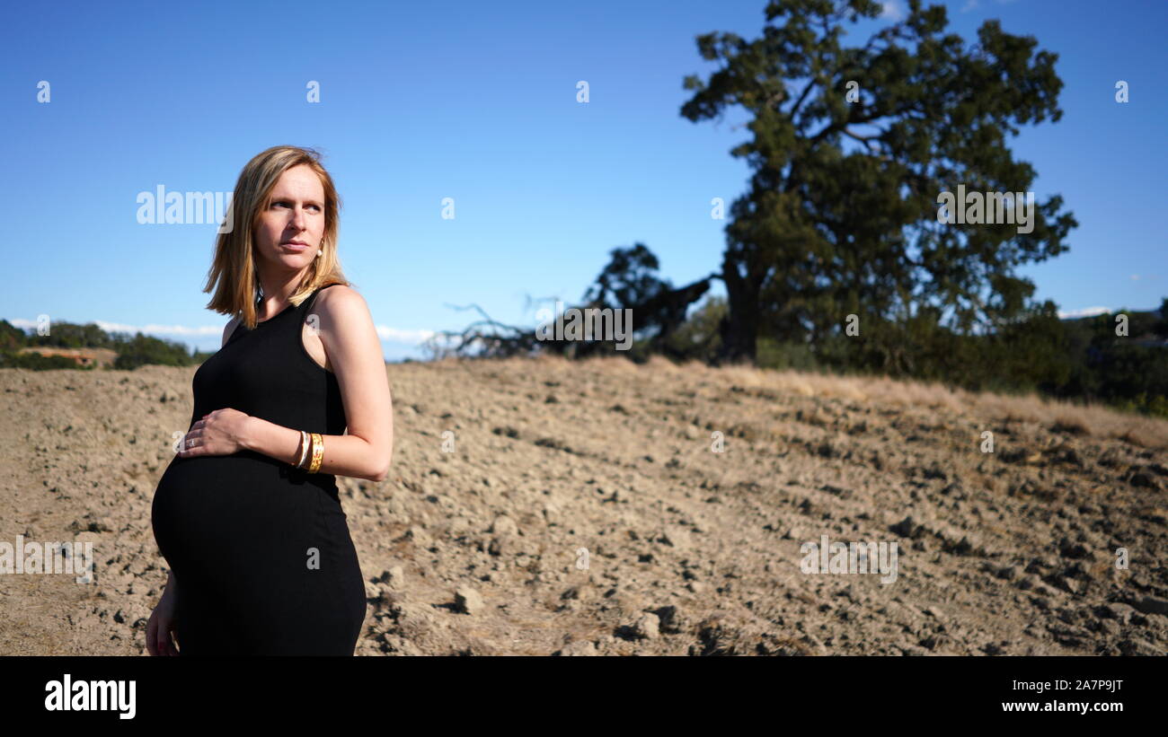 Young blonde woman wearing stretchy black dress, obviously very pregnant, standing in foreground of rural country scene of dirt ground and large tree Stock Photo