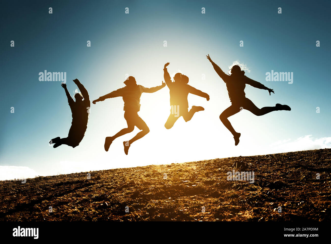 Four silhouettes of friends are jumping together against blue sky and sun Stock Photo