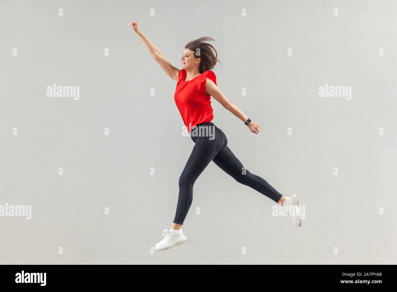 Full lenght of happy beautiful brunette young woman in casual red shirt jumping up in super hero pose and looking forward with toothy smile. indoor, s Stock Photo