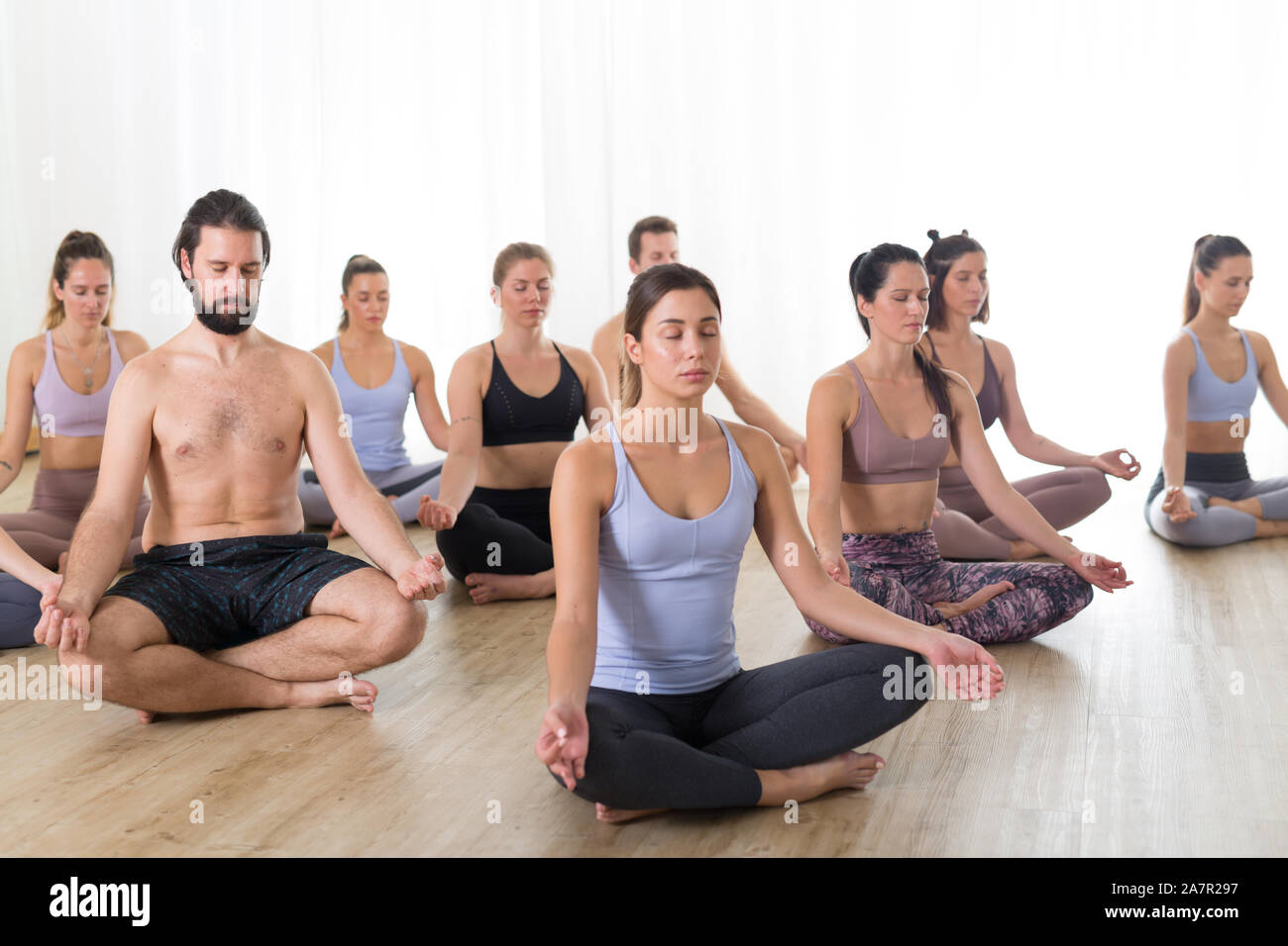 Group of young sporty attractive people in yoga studio, practicing yoga lesson with instructor, sitting on floor in Siddhasana, easy seated yoga pose Stock Photo