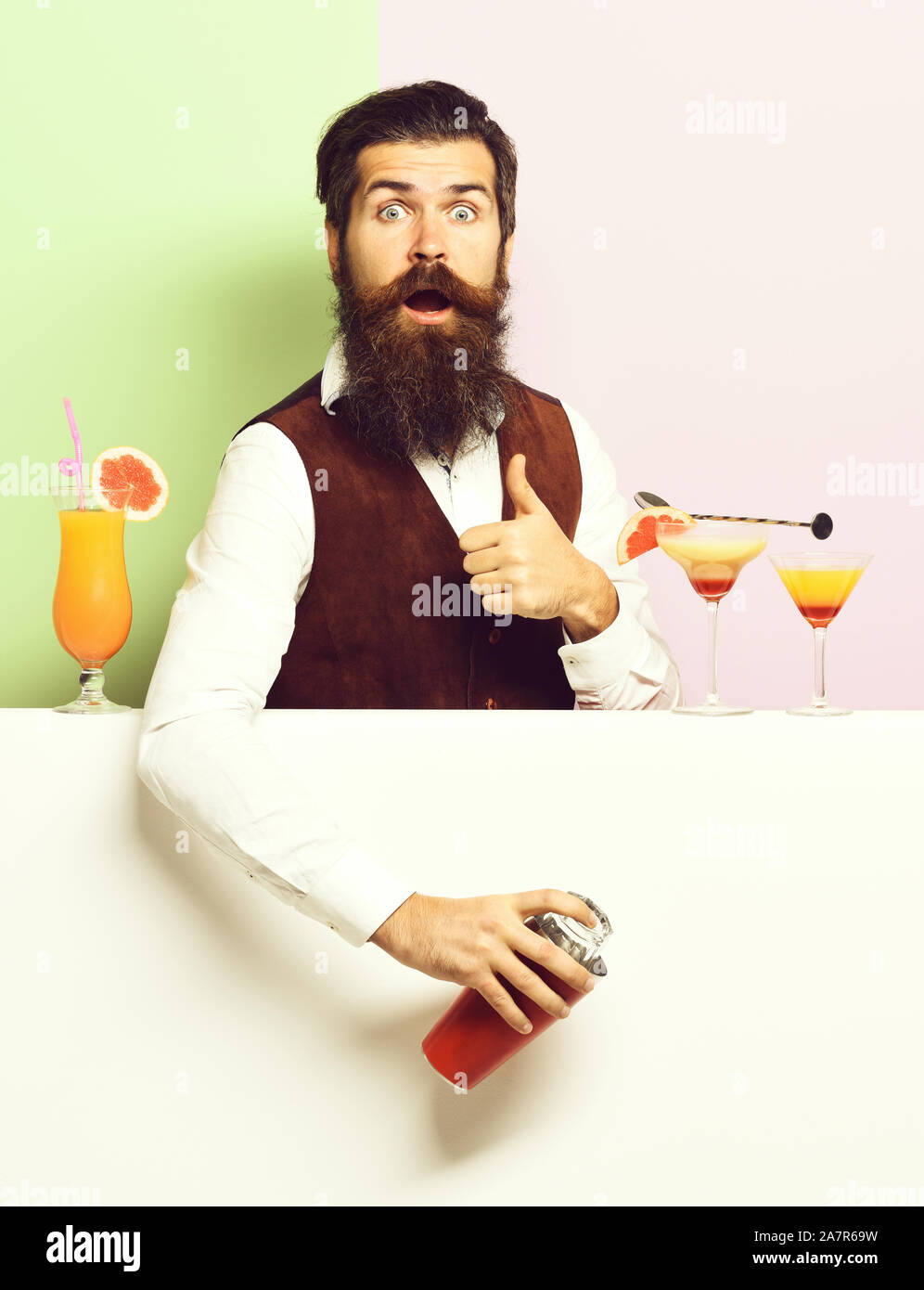 handsome bearded barman with long beard and mustache has stylish hair on surprised face holding shaker and made alcoholic cocktail, in vintage suede leather waistcoat on purple green studio background Stock Photo