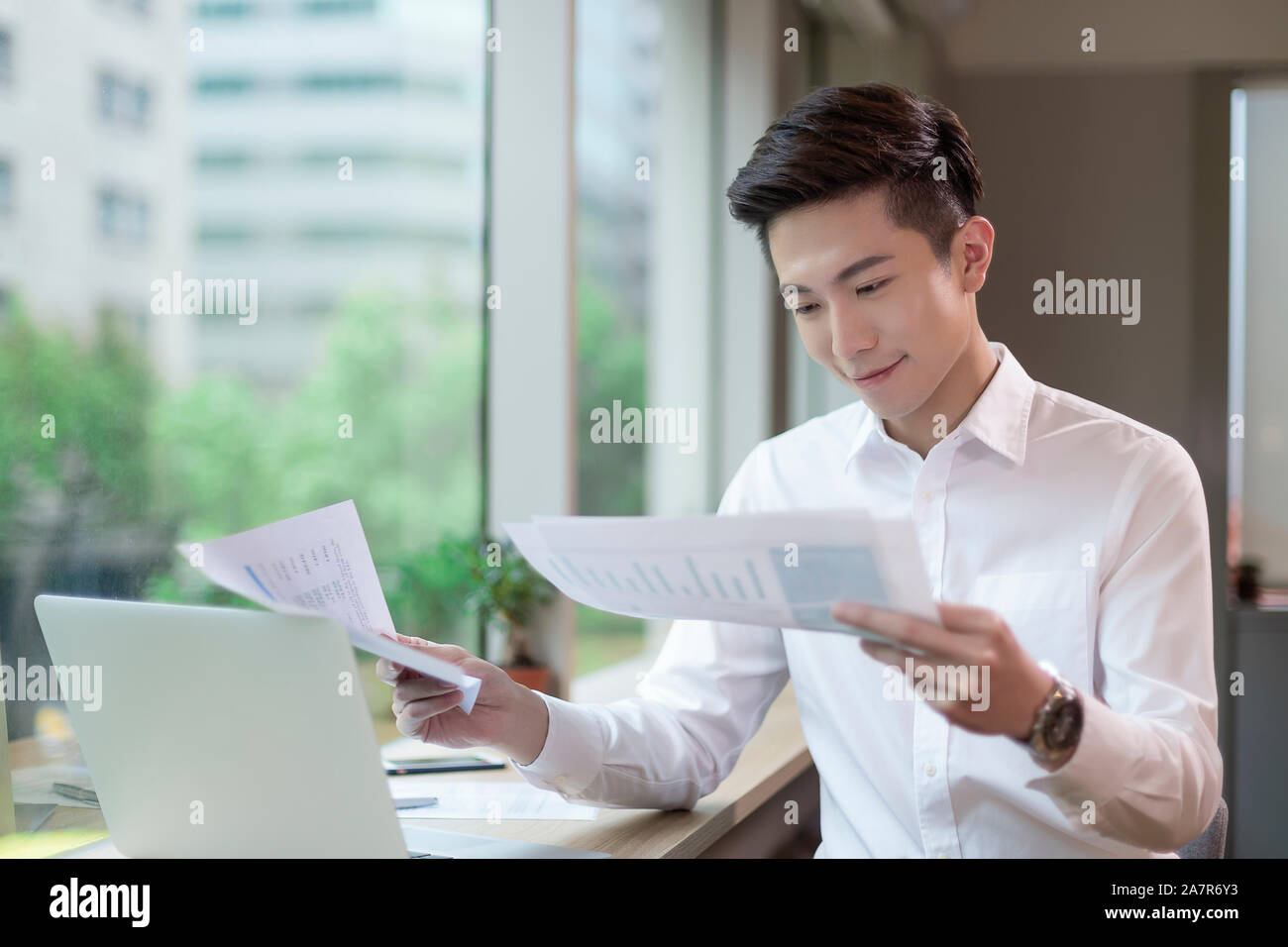 Waist up shot of a busy smiling young businessman with short black hair and a white button down reading documents at a desk in an office Stock Photo