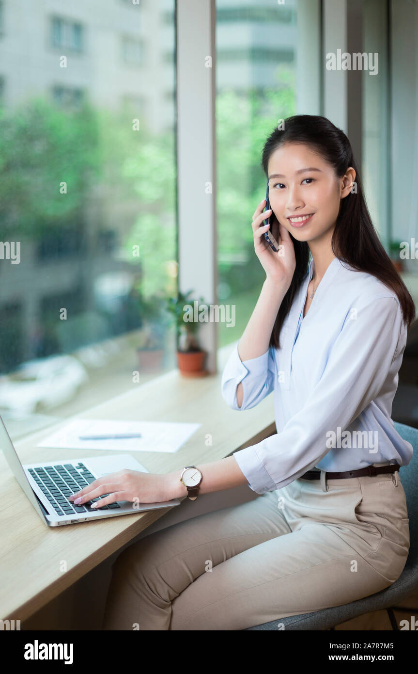 Smiling young businesswoman with long straight black hair using a smartphone and a laptop while looking at the camera at a desk in an office Stock Photo