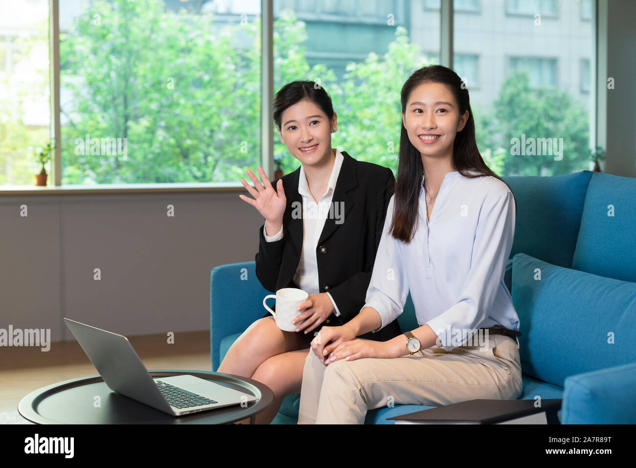Photograph of two young businesswomen sitting on a blue sofa in an office and smiling at the camera with one also waving Stock Photo
