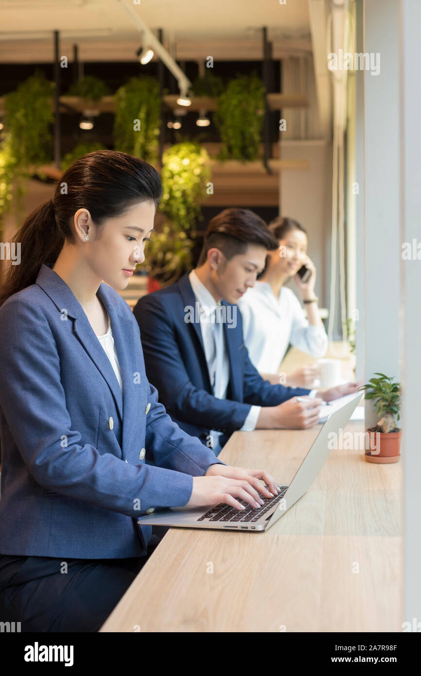 Group of three young male and female businesspeople working at a desk in an office with the woman in the foreground using a laptop Stock Photo