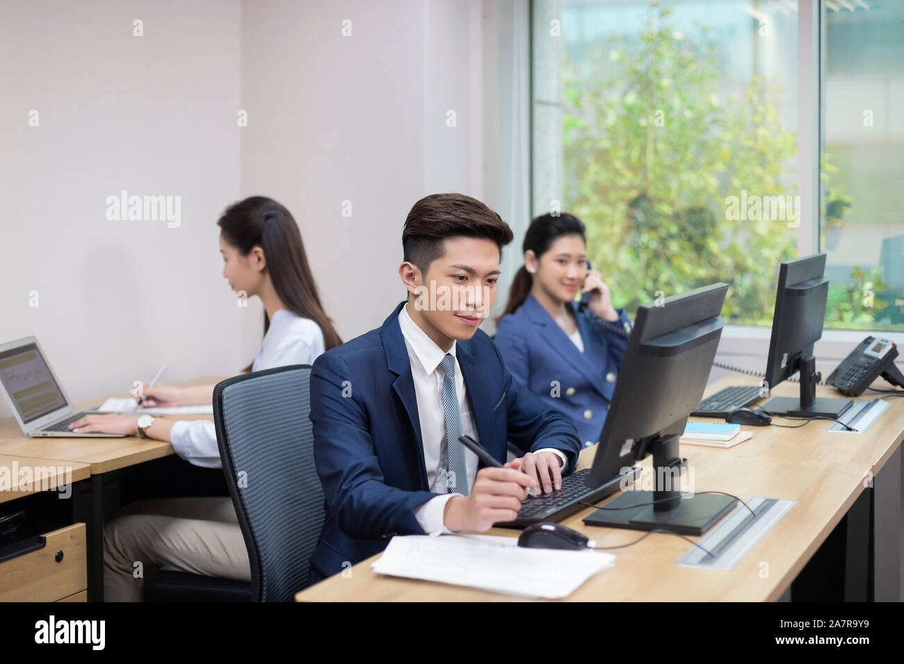 three young male and female businesspeople working in an office with one using computer, another one using phone and another one using laptop Stock Photo