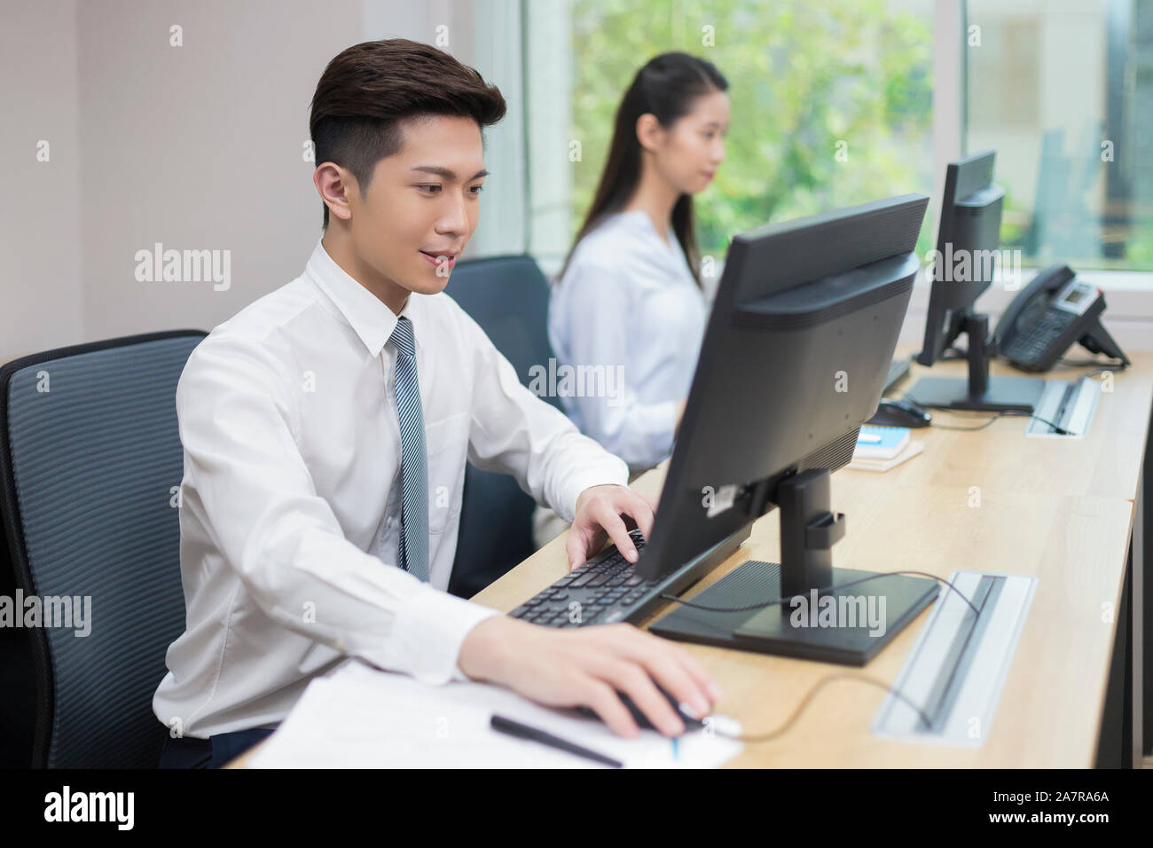 Side view waist up shot of a young businessman with black hair wearing shirt and a tie using a computer at a desk in an office Stock Photo