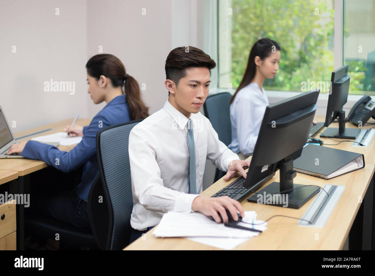 Photograph of three young male and female businesspeople working in an office with two using computers and another one using laptop Stock Photo