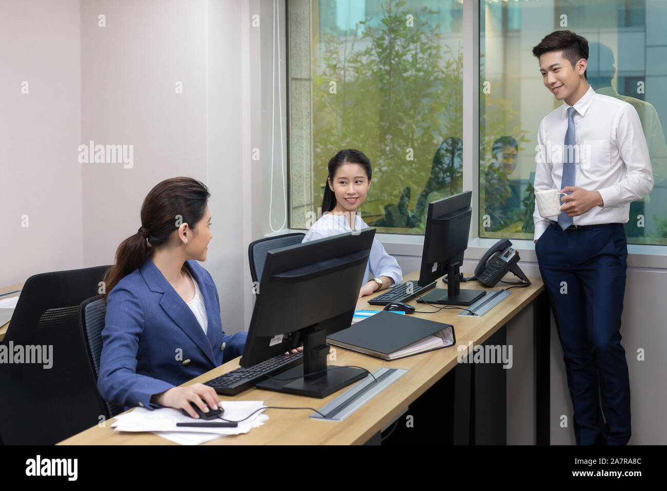 Photograph of three male and female businesspeople in an office with two of them talking while using computers at their desks and another one standing Stock Photo
