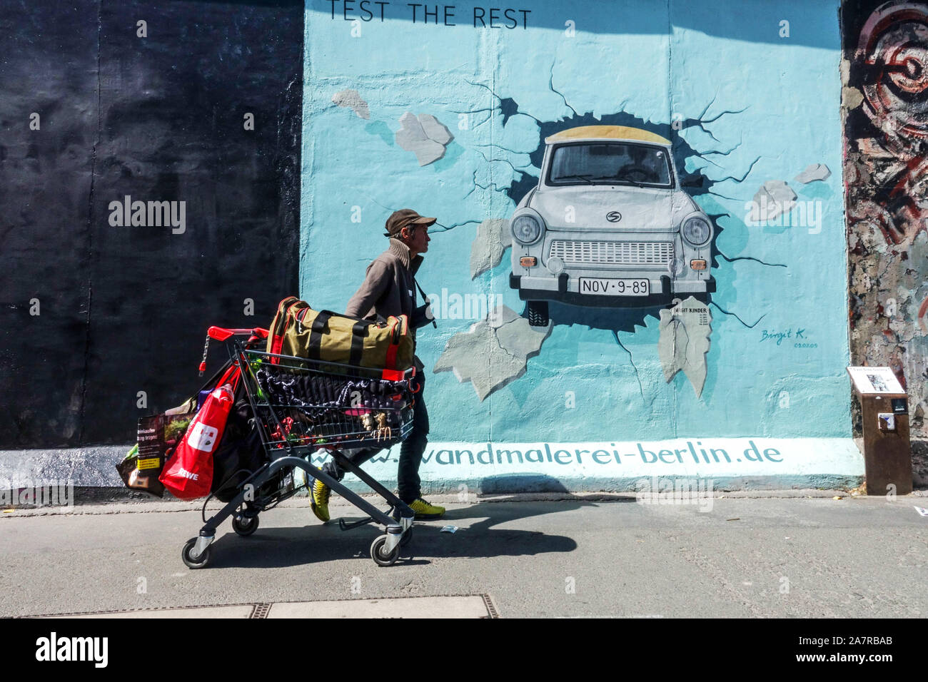 Berlin wall graffiti Migrant Homeless or Economic tourist with supermarket trolley passing around Trabant car in East Side Gallery Germany Stock Photo