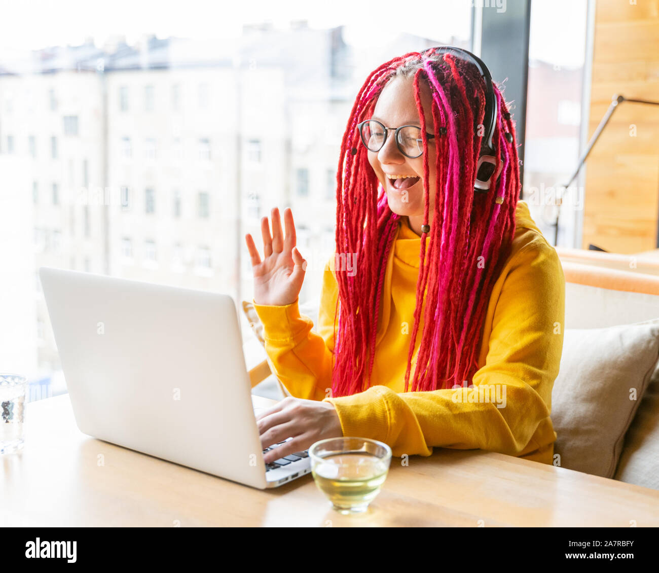 The woman in headphones looks at the laptop, waves her hand and smiles. Live chat, stream. Blogger girl with long pink hair. Good luck concept, positi Stock Photo