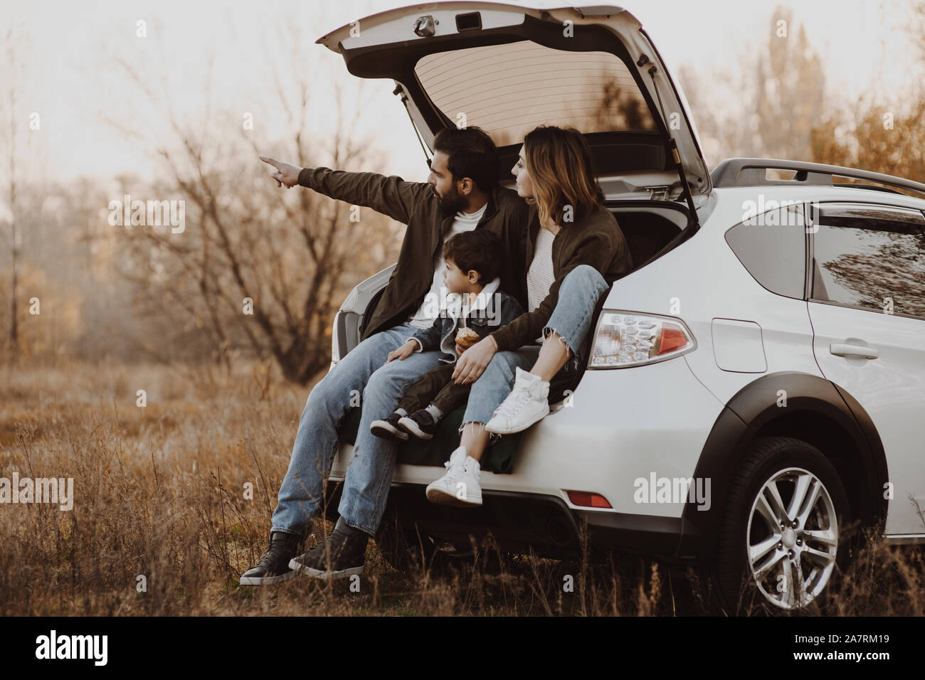 Happy family sitting at open trunk of hatchback car and pointing finger outdoors. Road trip concept. Stock Photo