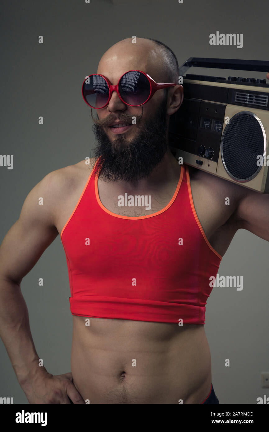 Portrait of a bearded man in black glasses and a tight fitness t-shirt holding a tape recorder on his shoulder. Preparing for a retro party Stock Photo