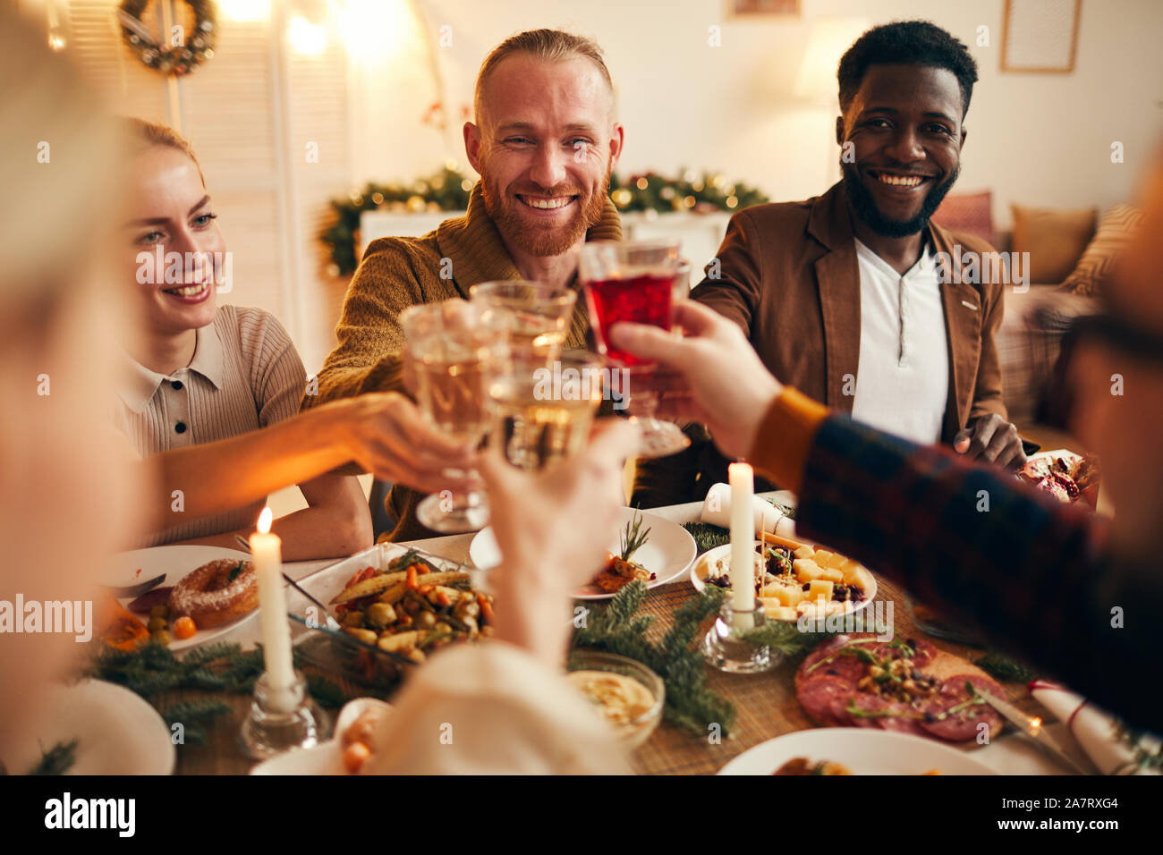 Multi-ethnic group of cheerful adult people clinking champagne glasses while enjoying Christmas dinner at home, copy space Stock Photo