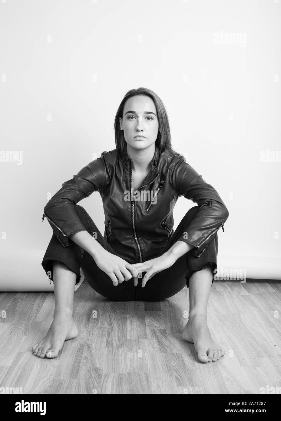 Black and white portrait of a barefoot young woman wearing a leather jacket and sitting on floor Stock Photo