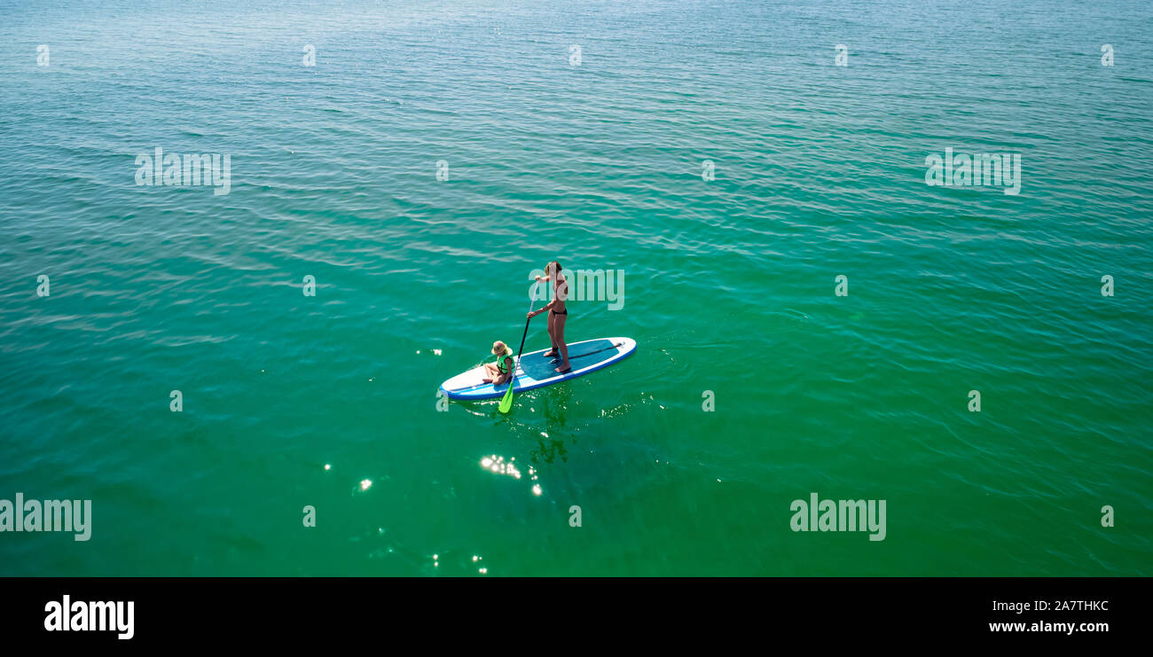 Little girl in a life vest sitting on the paddle board with mother