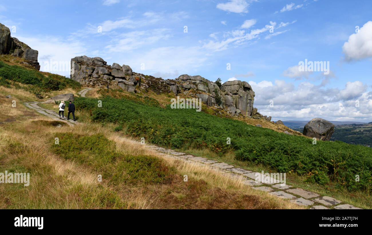 People walking on path to high sunlit rocky outcrop under blue sky - scenic landscape of Cow and Calf Rocks, Ilkley Moor, West Yorkshire, England, UK. Stock Photo