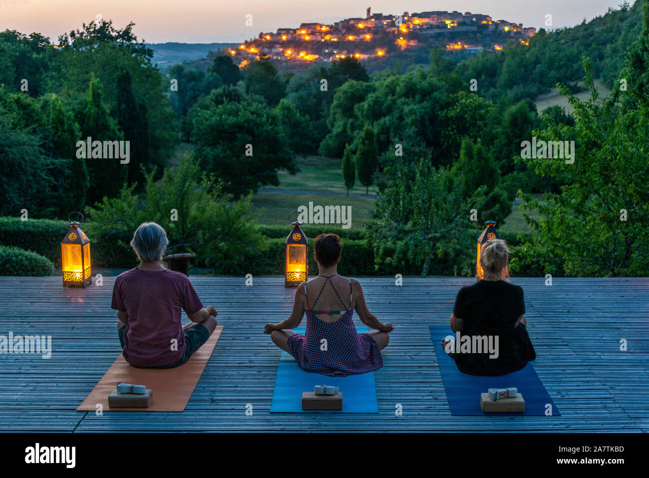 Three people practicing yoga at dusk,on a wooden platform, overlooking the medieval village of Cordes-sur-Ciel, in Occitanie, France. Stock Photo