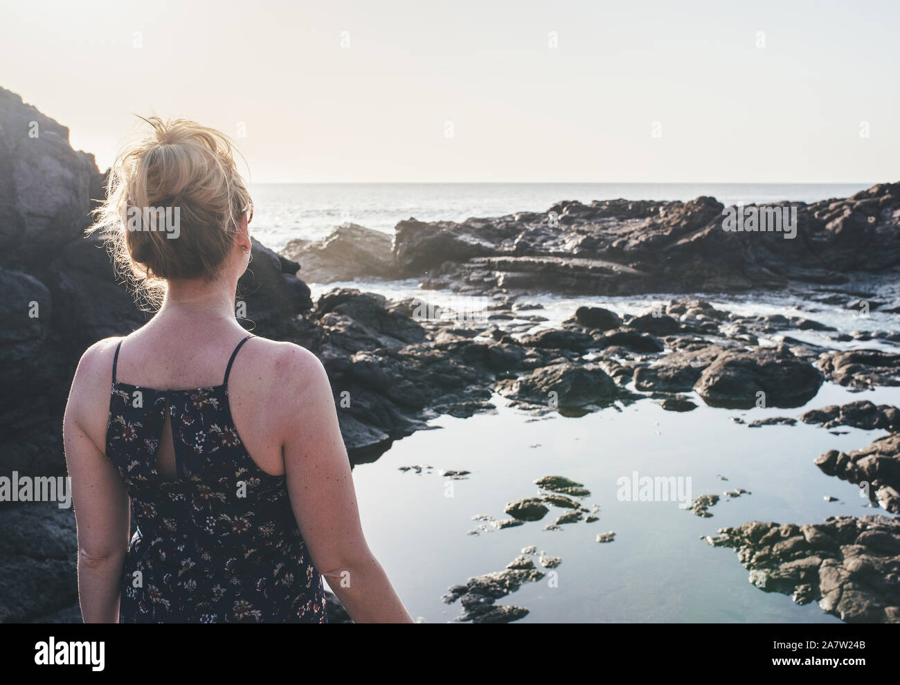 blonde woman in summer dress standing at rocky shore looking at ocean in sunlight Stock Photo