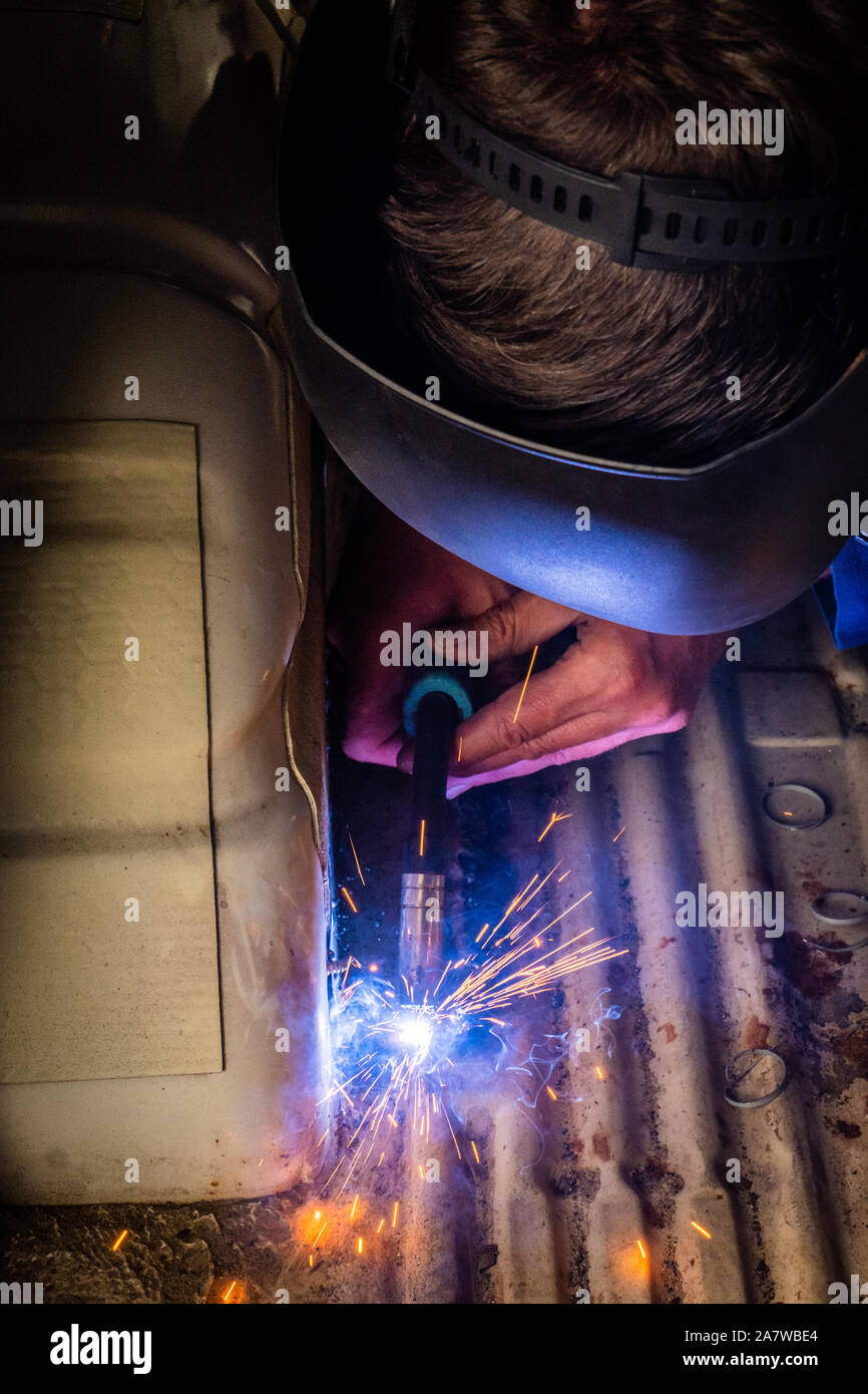 Man with a helmet and no gloves welds in the metal of a car and repairs a rusty hole Stock Photo