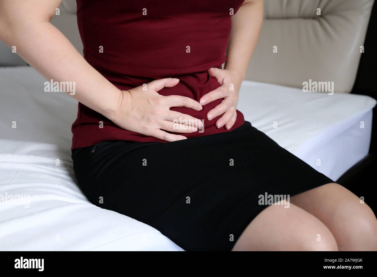 Woman suffering from pain in abdomen. Slim girl in black skirt sitting on bed clutching her belly, concept of indigestion, stomach ache, menstruation Stock Photo