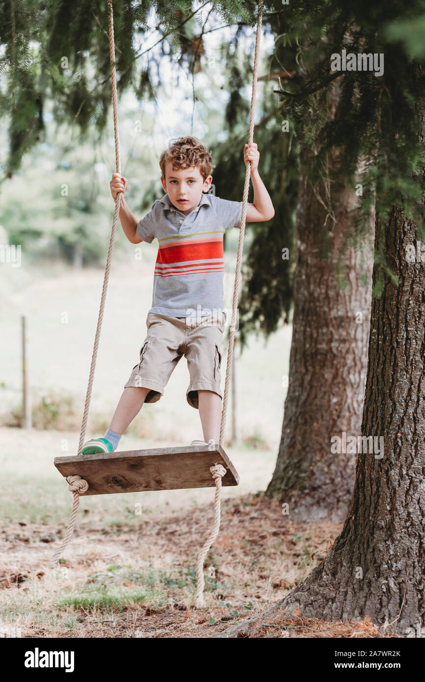 Vertical front view of boy standing on swing under pine trees Stock Photo