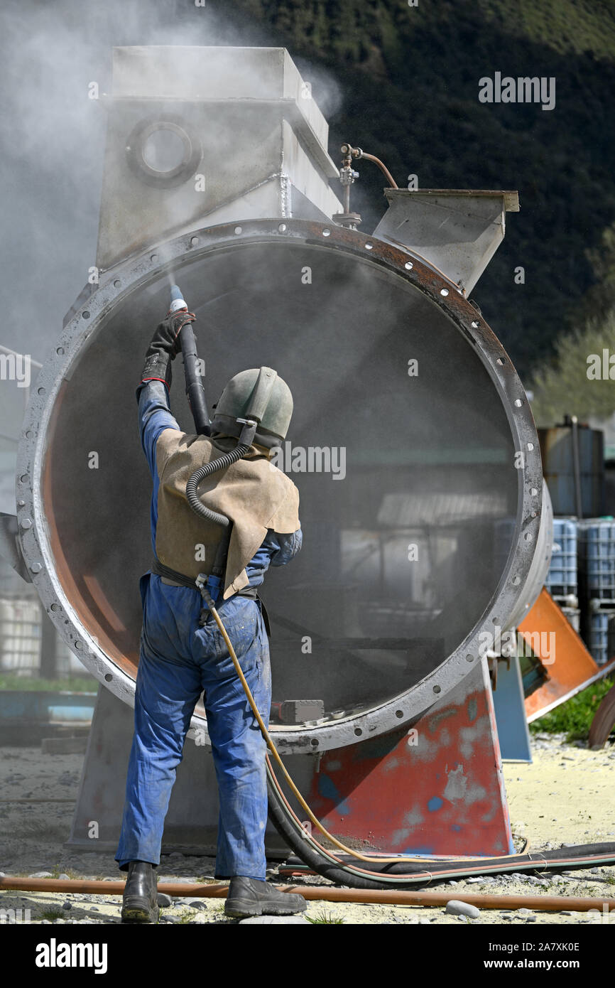 A man weating full safety gear uses ground glass to sandblast the steel casing of a meat drying machine for a rendering plant Stock Photo