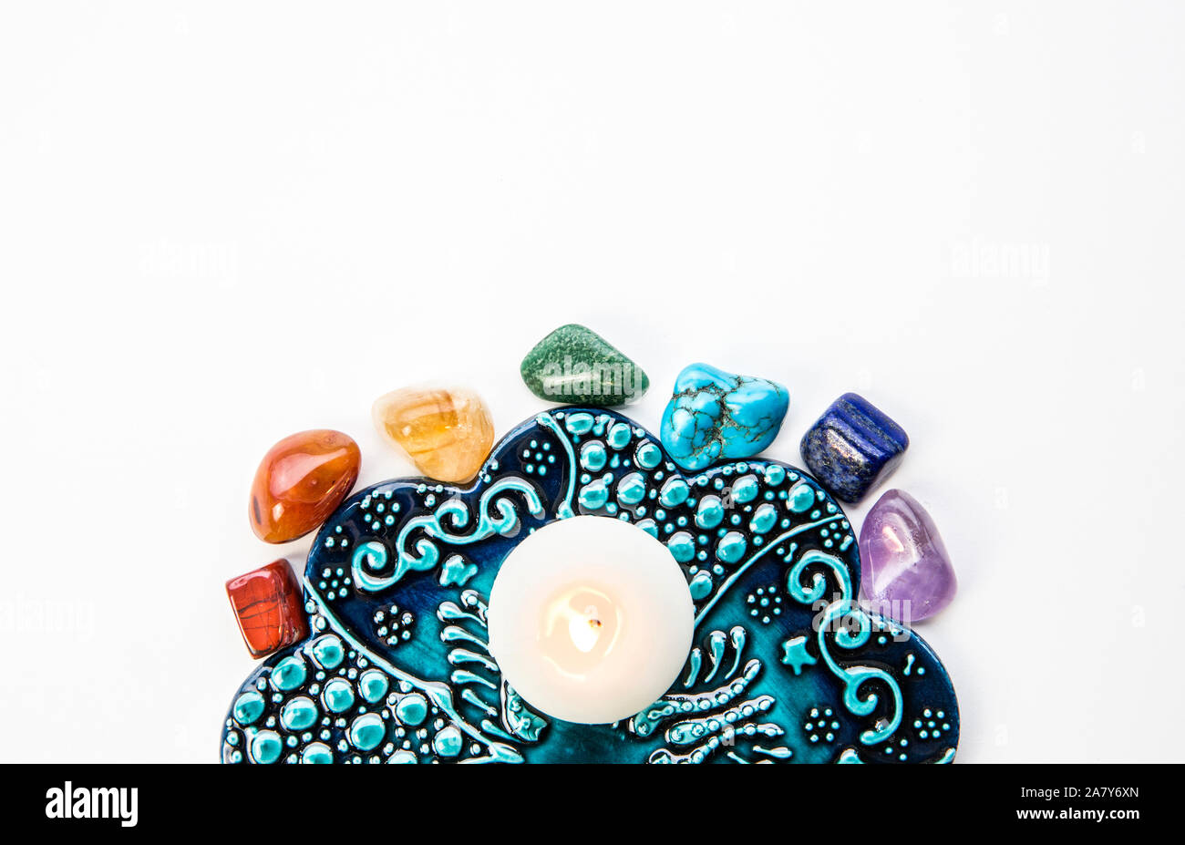 Top view of all 7 chakra colors semi precious crystal stones on blue and white ornament background with relaxing candle burning in center. Lot of copy Stock Photo