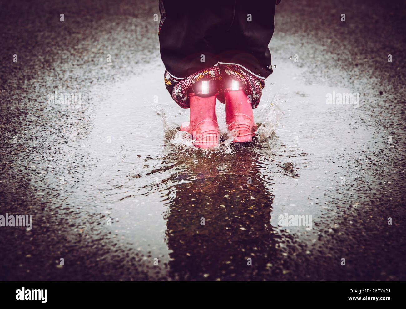Girl having fun, jumping in water puddle on wet street, wearing rain boots with reflective detail fabric stripes shining. High visibility and safety i Stock Photo