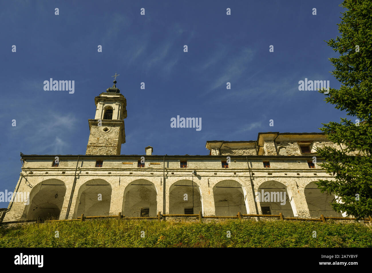 Low angle view of the Sanctuary of St Magno with an ancient arcade and the stone bell tower against clear blue sky, Castelmagno, Piedmont, Italy Stock Photo