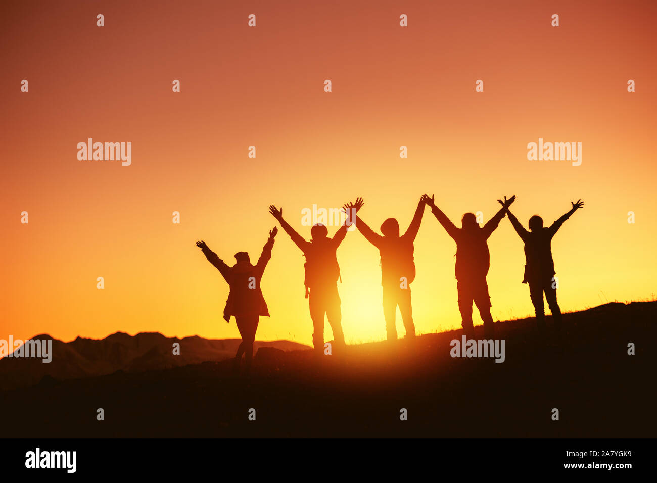 Group of happy peoples silhouettes stands with raised arms against sunset and mountains Stock Photo