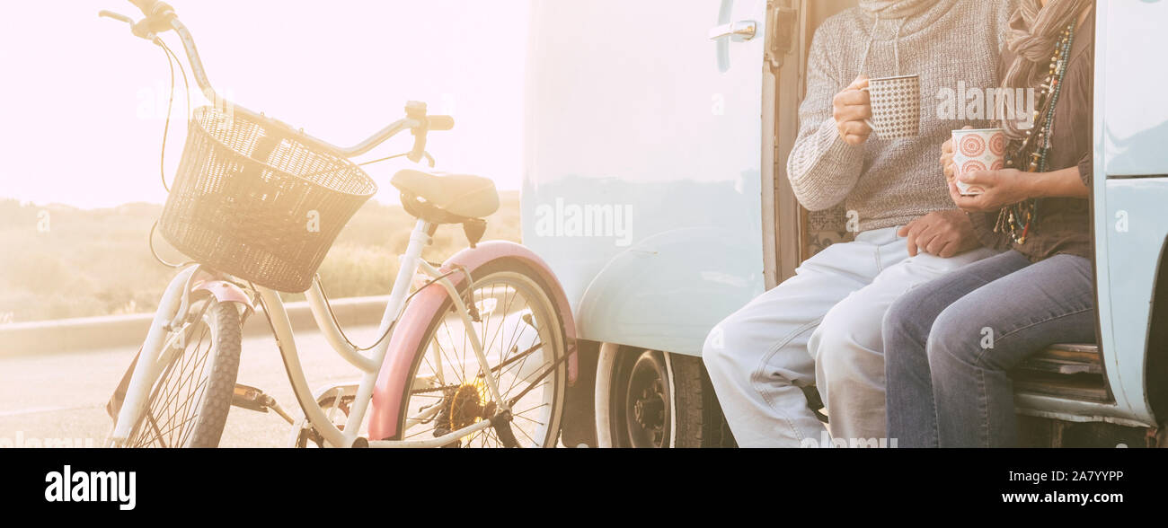 Unrecognizable adult senior couple sitting on an old vintage van together drinking tea or coffee - old bikes and sunlight in background - concept of l Stock Photo