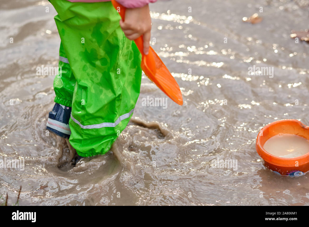 Low section of a child with rubber boots and waterproof pants playing in a deep muddy rain puddle with a shovel and a bucket. Seen in Bavaria in Germa Stock Photo