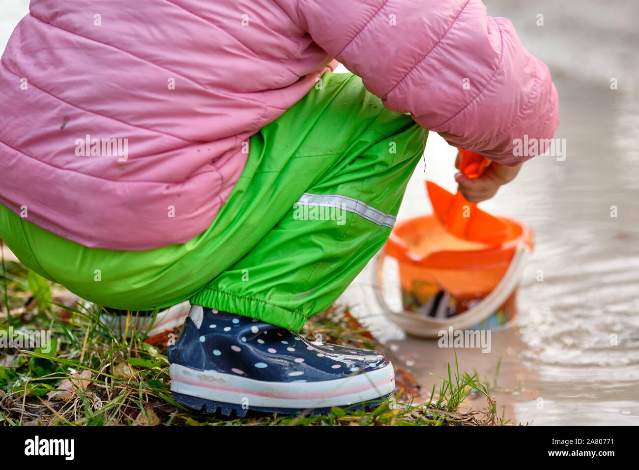 Low section of a child with rubber boots and waterproof pants playing in a deep muddy rain puddle with a shovel and a bucket. Seen in Bavaria in Germa Stock Photo