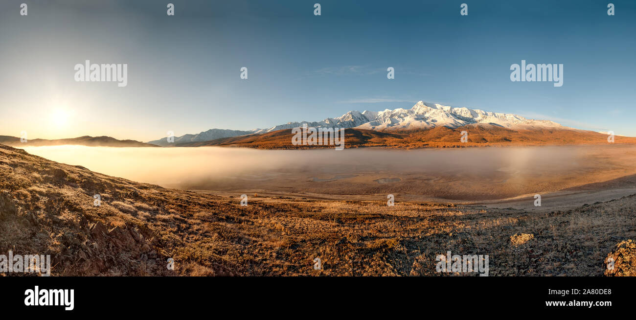 Amazing autumn panorama with the sun, mountains covered with snow and a golden forest, white fluffy fog over the lakes and the valley against the blue Stock Photo