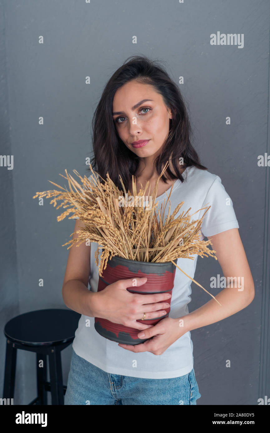 Beautiful Caucasian girl with short hair holding a vase with wheat ears standing on a gray background Stock Photo