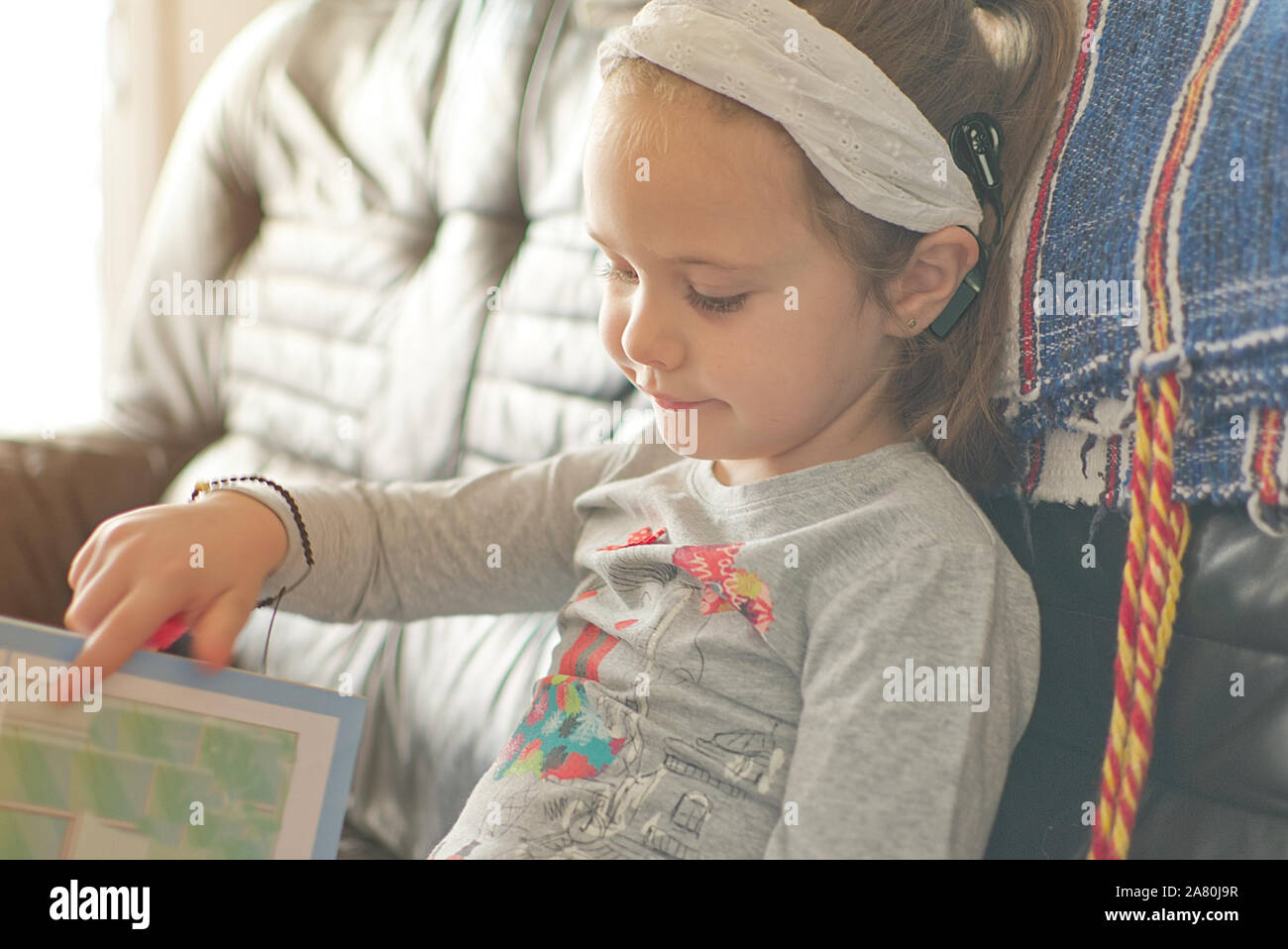 Blonde little girl with cochlear implant reading a book at home. Hear impairment and deaf community concept with empty copy space for Editor's text. Stock Photo