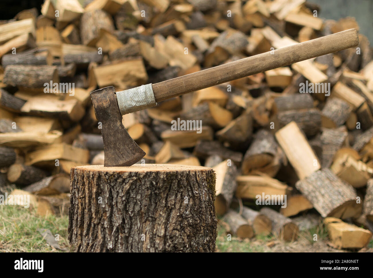 An axe stuck in a log in front of a pile of wood. Stock Photo