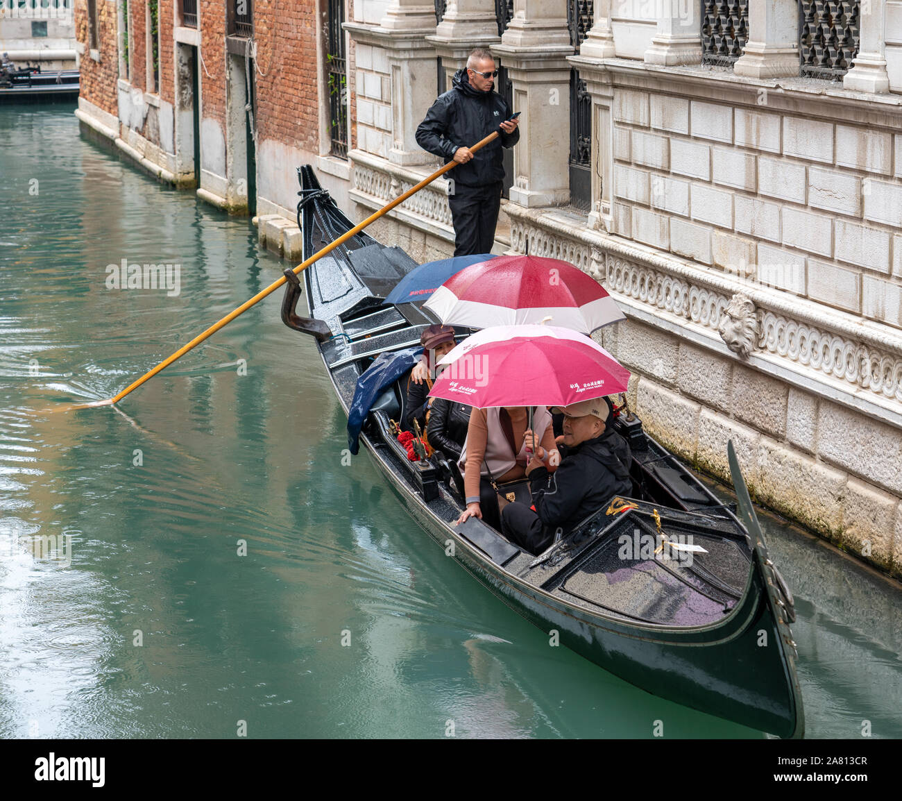 A gondolier looking at his mobile phone while ferrying tourists around a rainy Venice Italy Stock Photo