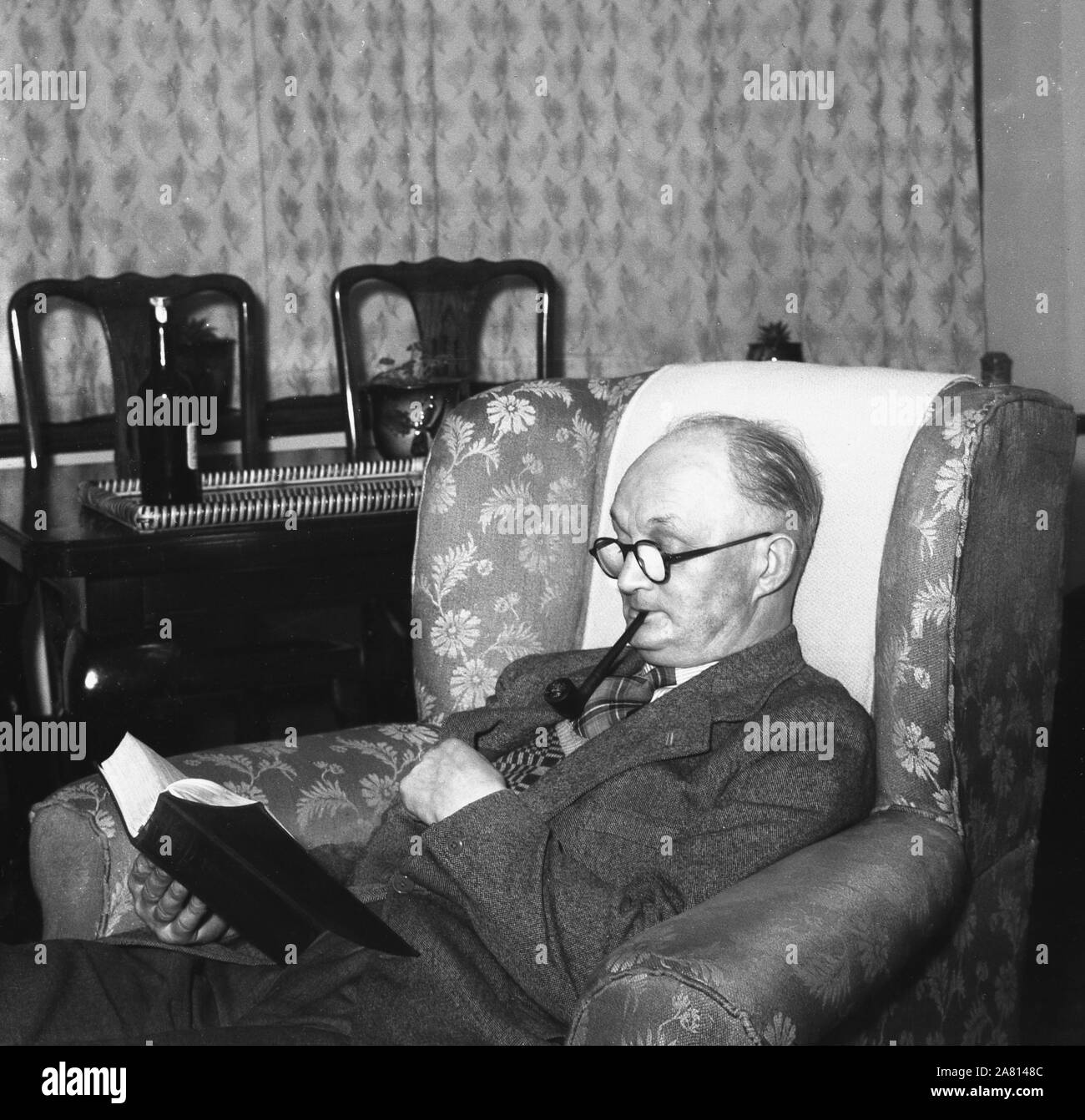 1950s, historical, an elderly man in a suit & tie, sitting in an armchair smoking a pipe and reading a book, England, UK. Stock Photo