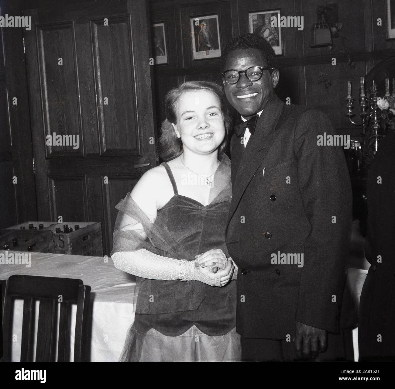1950s, historical, posing for a photo at a black tie dance, a young English lady with a young African man, England, UK. Post-war Britain saw many people from the Commonwealth countries arriving in Britain to work and study and cultural exchanges and dances were held to introduce them to the British way of life. Stock Photo