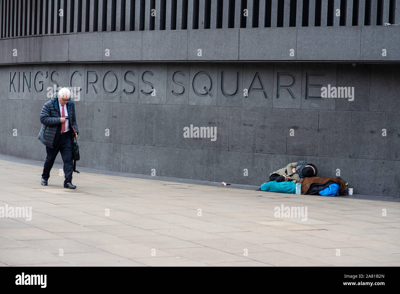 London, United Kingdom - November 5, 2019: A man in a suit walks through King's Cross Square while a rough sleeper rests on the pavement. Stock Photo