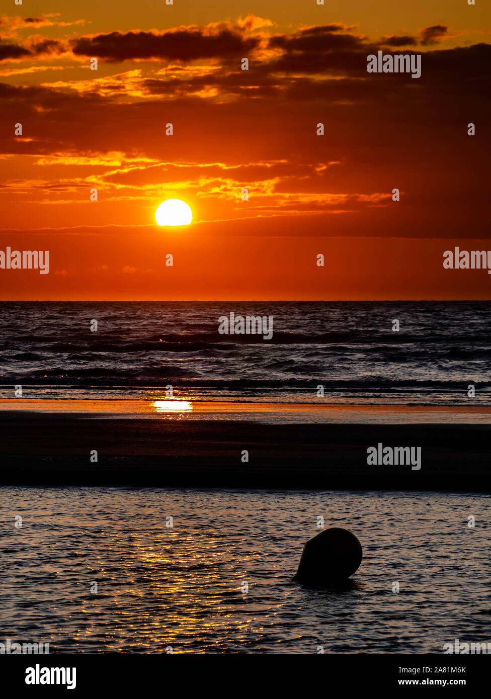 Colorful sunset at the beach with the sun peeking below a cloudlayer and a buoy at the foreground Stock Photo