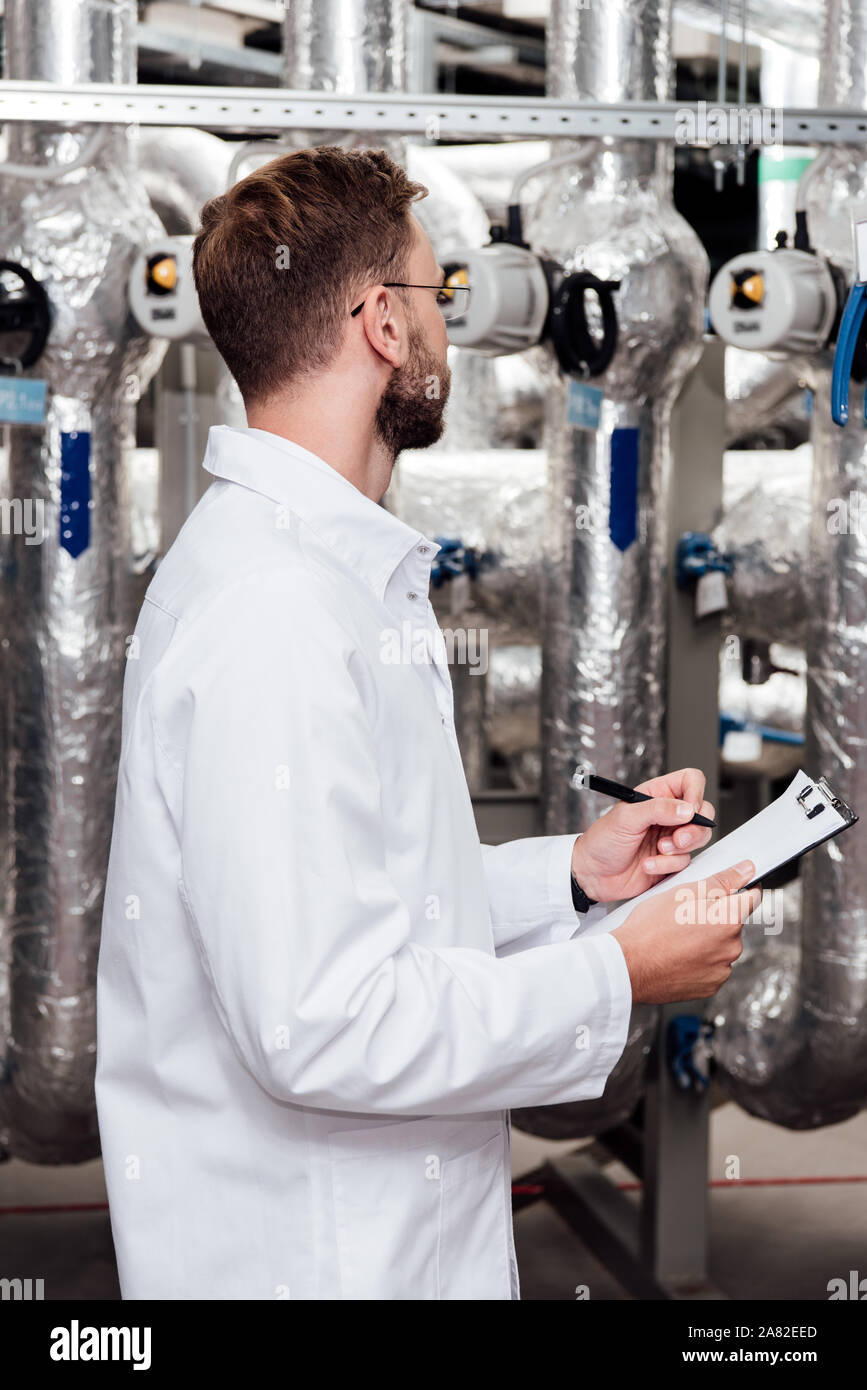 bearded engineer in white coat holding clipboard and pen near air compressed system Stock Photo