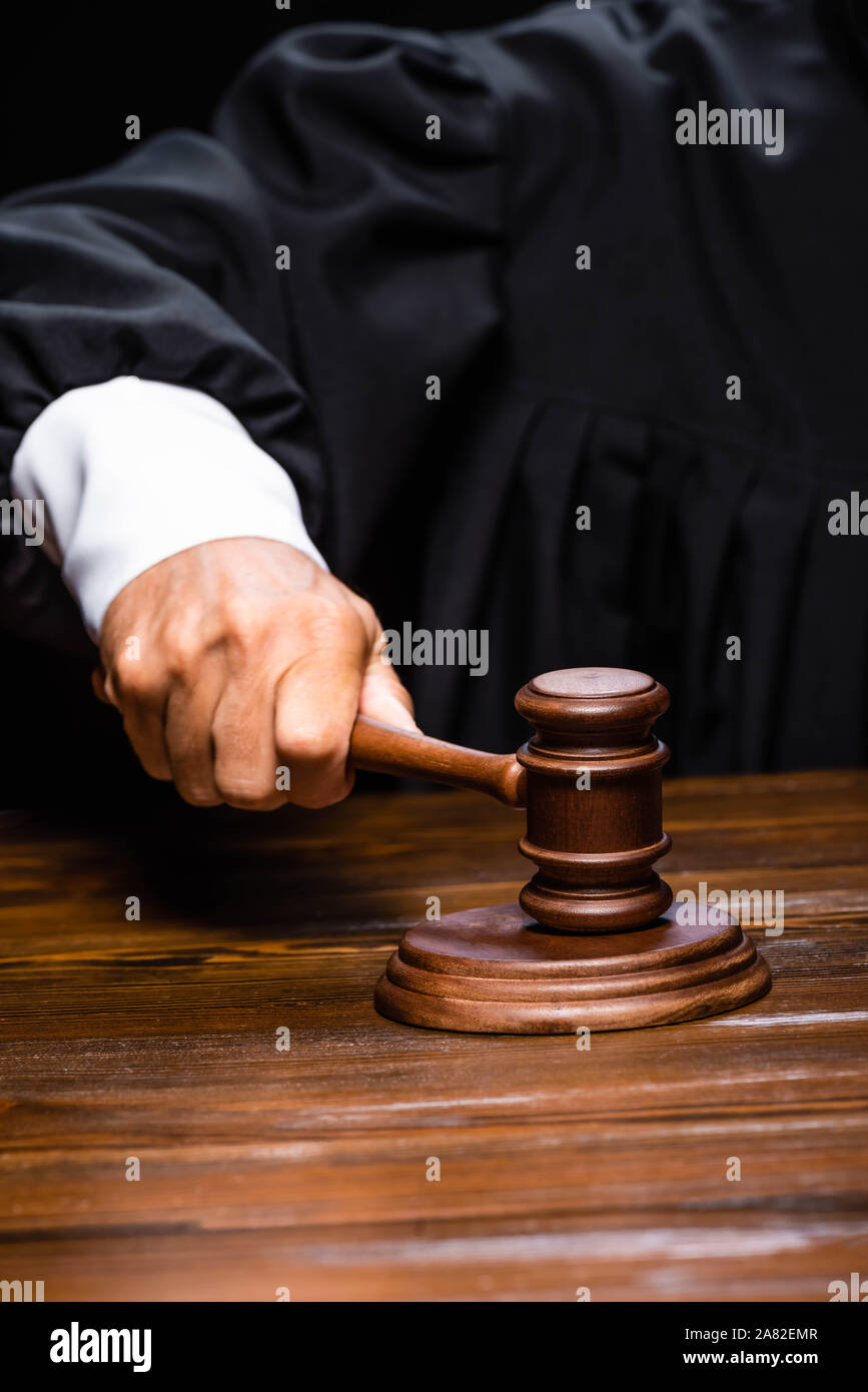 cropped view of judge in judicial robe sitting at table and hitting with gavel isolated on black Stock Photo