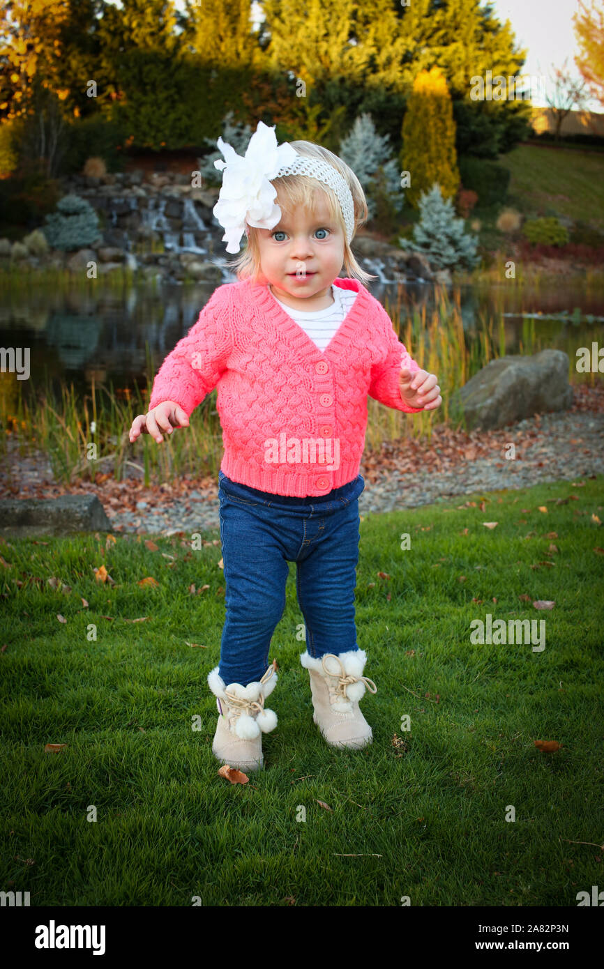 Very Cute Toddler Blonde Girl with Big Blue eyes Smiling in the middle of the Beautiful Autumn Park wearing Hot Pink Jacket and Blue Skinny Jeans and Stock Photo