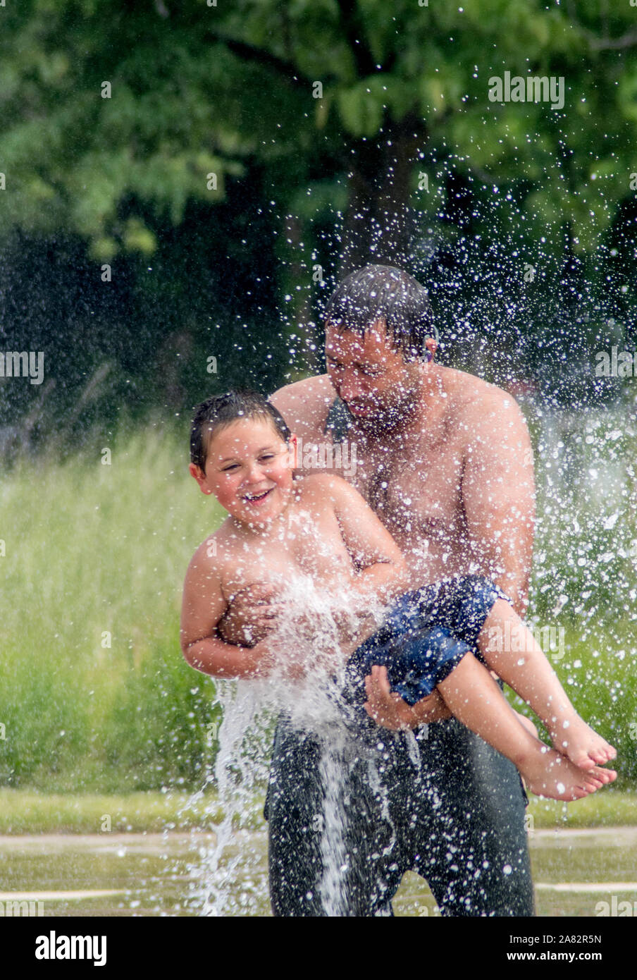 A father swings his young son through a fountain at a local splash pad in Michigan USA Stock Photo