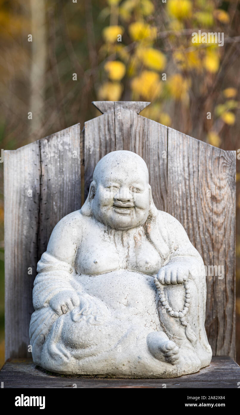 The Laughing Buddha, Budai, with his cloth sack stone sculpture with yellow fall colors in the background in autumn Stock Photo