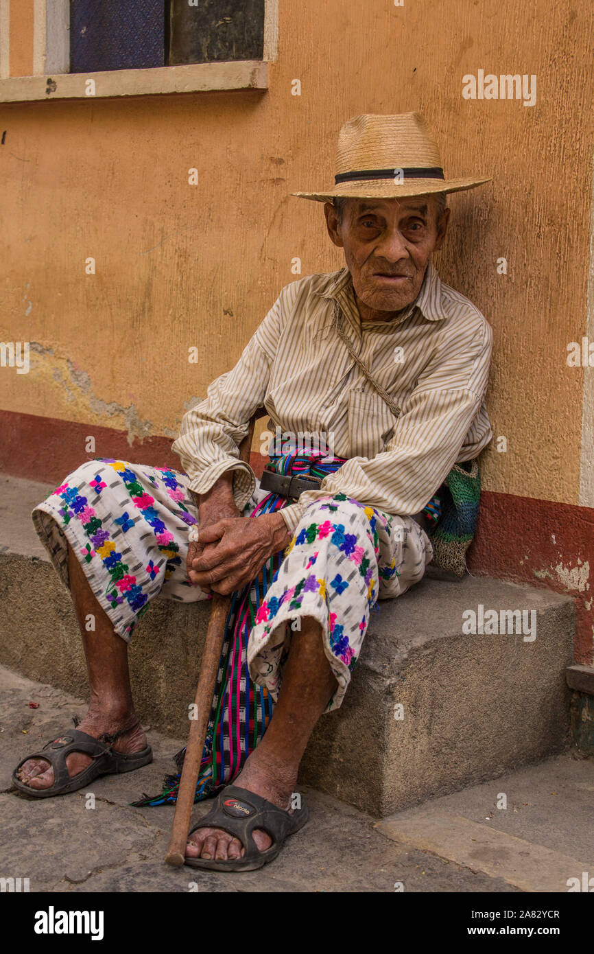 93 year old Mayan man in traditional dress in San Pedro la Laguna, Guatemala, sitting on the curb with a cane in front of a pastel wall. Stock Photo